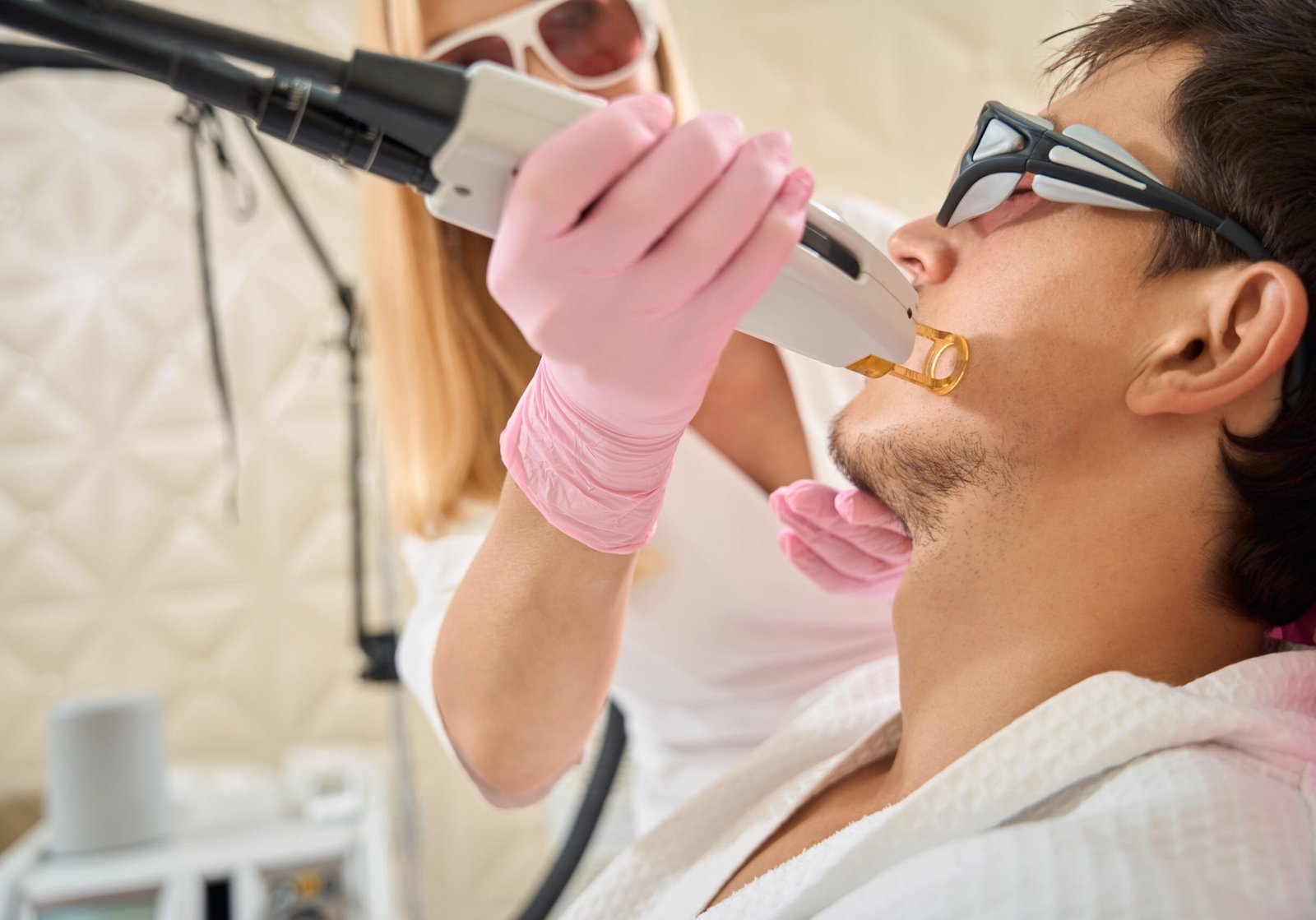 Female performs facial laser hair removal procedure on young client, people wearing safety glasses